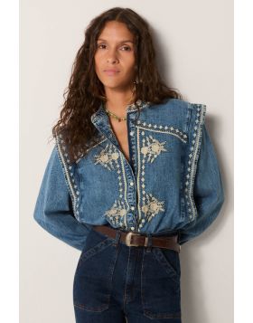 Cotton denim washed to structure yet softened by wear, carrying the quiet strength of workwear. Its surface feels steady, the faded blue closer to summer skies than indigo ink. Across chest and sleeves, ivory florals unfurl like stitched gardens, adding d
