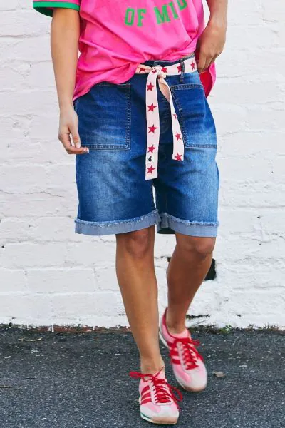 A pair of longer length darker denim stretch shorts with a relaxed fit. They have front pockets, a raw rolled hem, and sit mid-thigh. Styled with a tan belt, they offer a laid back, easy to wear beachy vibe. style H400.